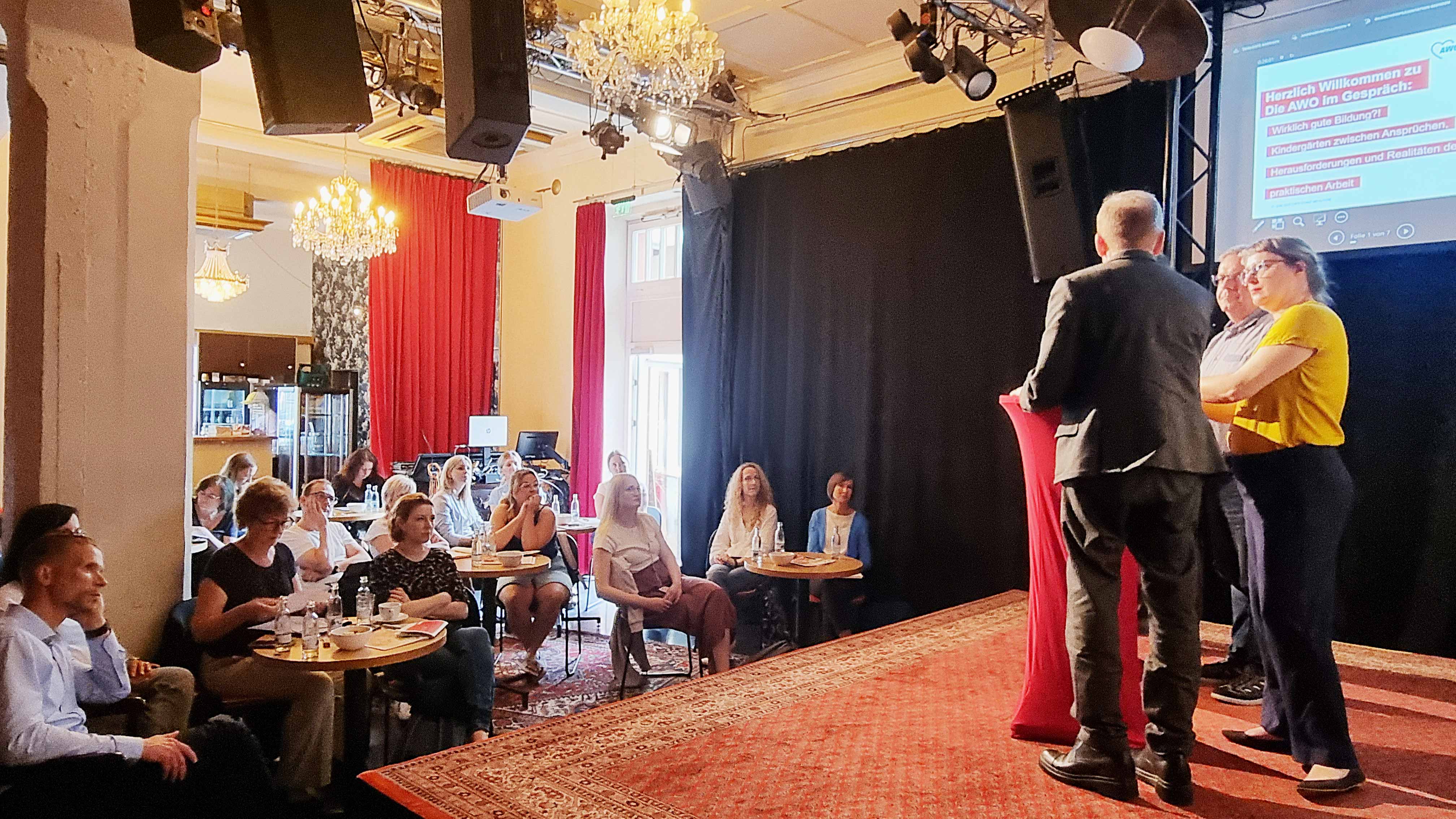 Zwei Männer und Ulrike Grosse-Röthig stehen am Stehtisch und unterhalten sich. Der Blick ist vn der Rückseite der Bühne Richtung Publikum, was in einer gemütlichen Atmosphäre an Tischen sitzt